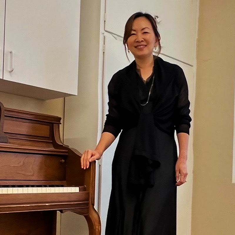 Singer Jenny Kim in a black dress posing next to an upright piano