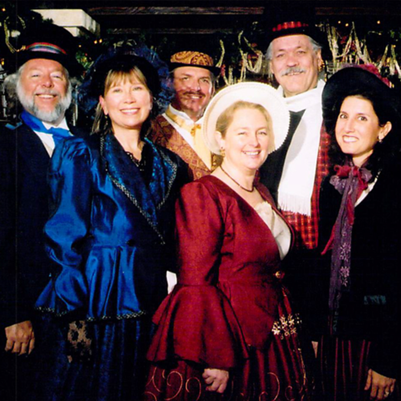 A group of 6 carolers in traditional Dickens costumes standing in two rows.