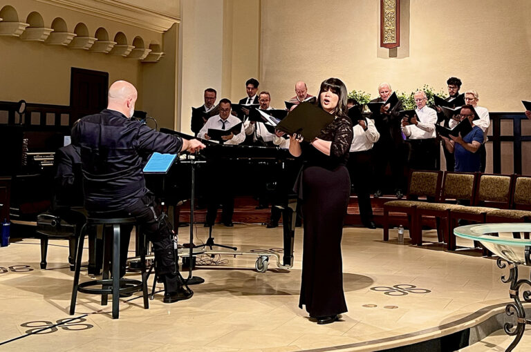 A female soloist holding a music folder sings in front of a male chorus