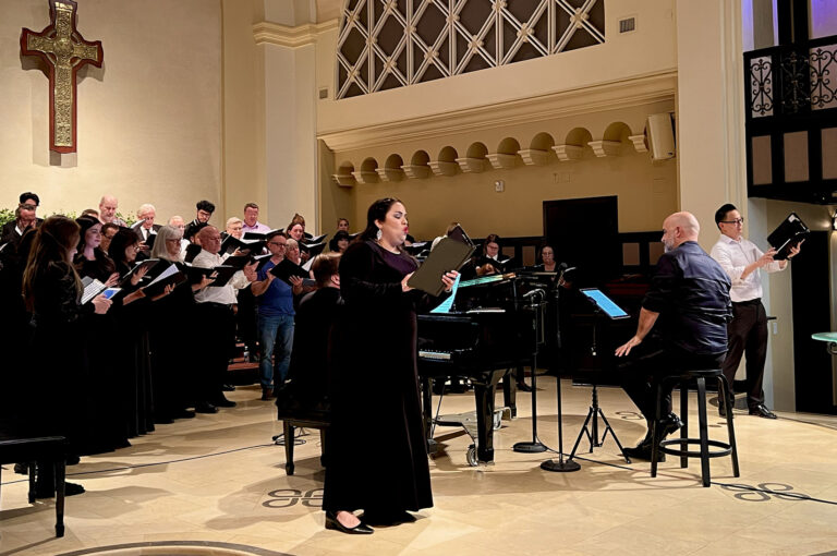 A male and female singer stand on either side of the conductor in front of a choir rehearsing.