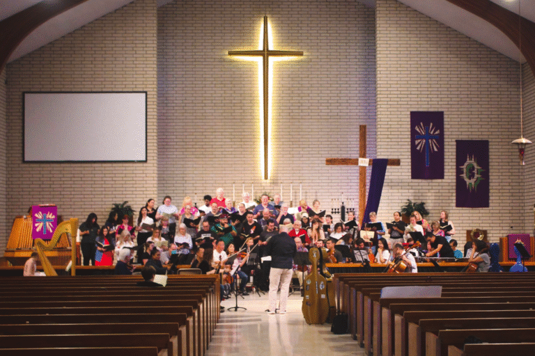 The choir and orchestra rehearsing on the alter of a church with a large lighted cross.