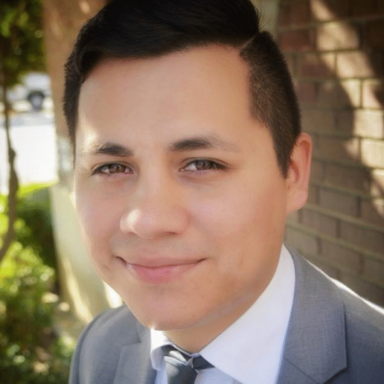 Headshot of a Male in a grey suit in a nature setting and a brick wall.