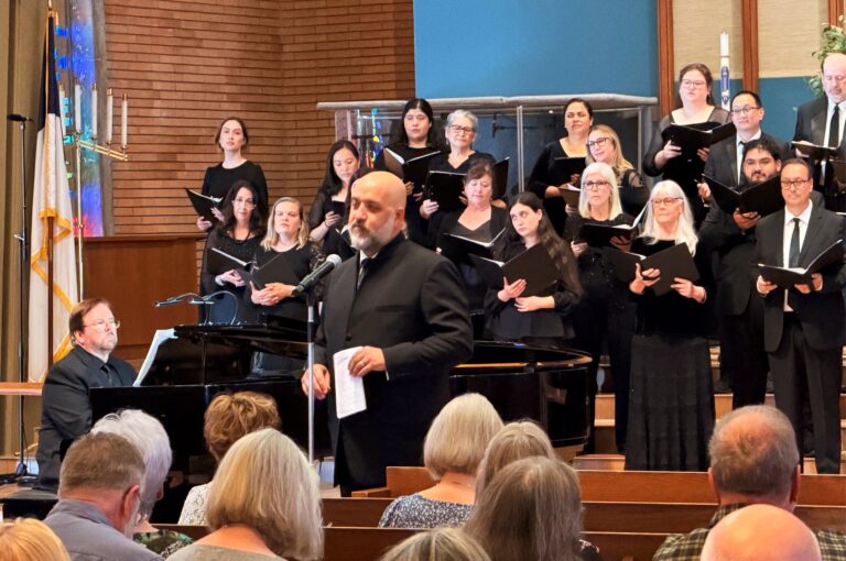 Conductor stands in front of a choir, dressed in black, and talks to the audience.