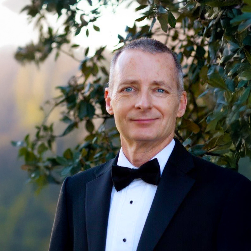 Guest conductor, Todd Helm in a black tuxedo standing in front of a tree.